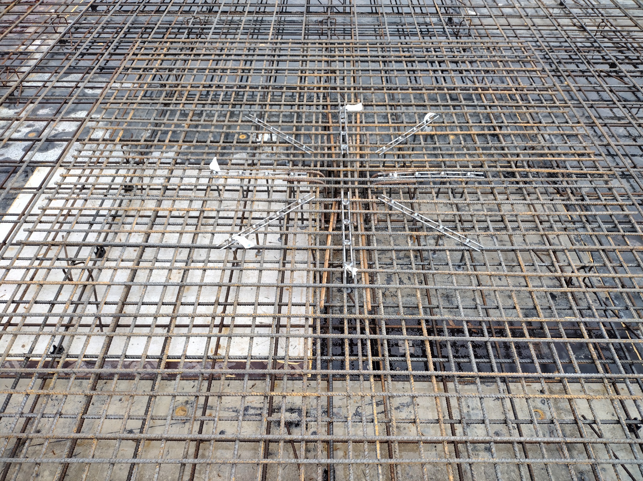 Siatka zbrojeniowa na fundamencie, widok z góry. Metalowe pręty tworzą regularną kratę, wzmocnioną w centralnej części dodatkowymi elementami.