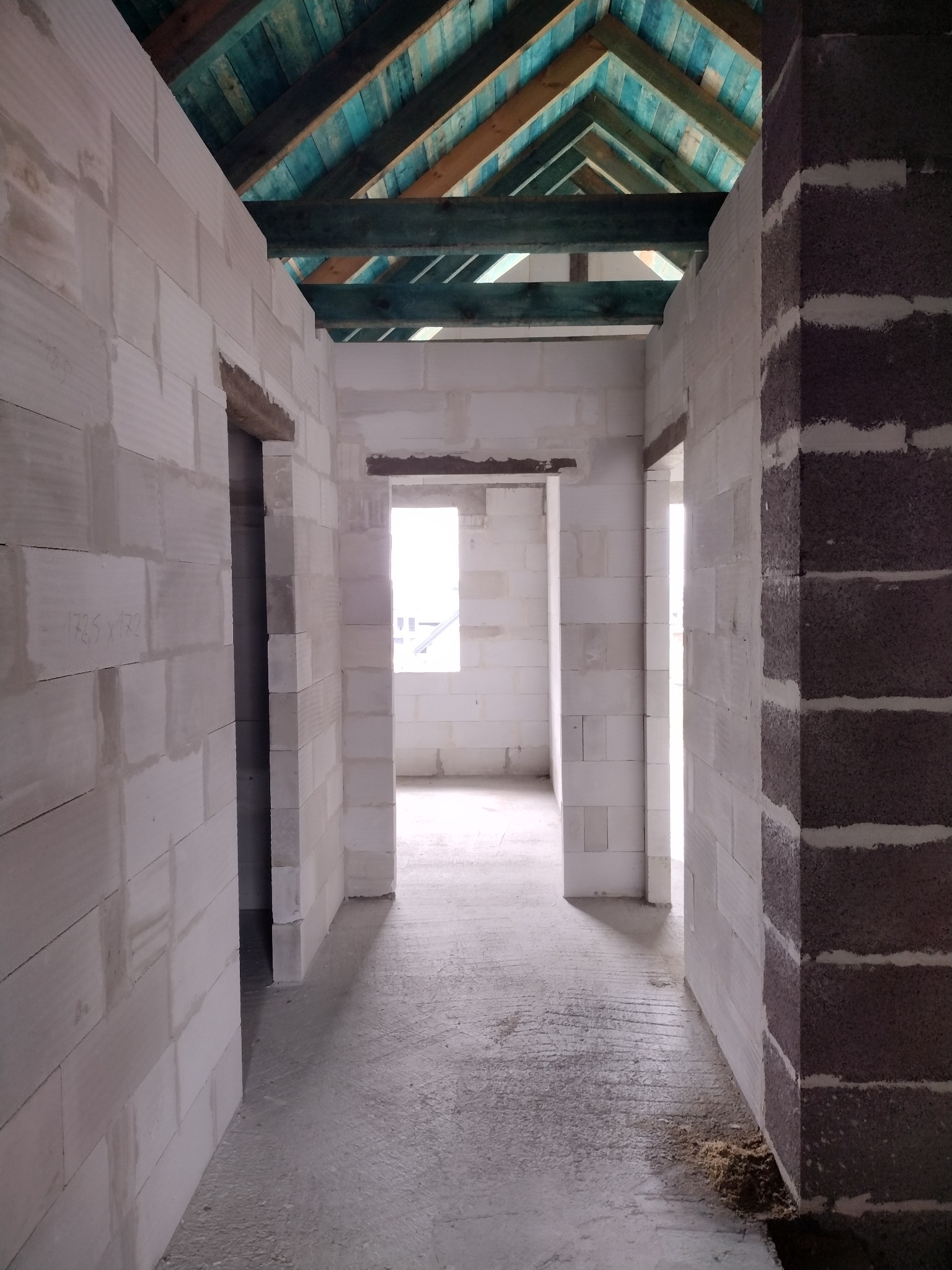 Wnętrze w trakcie budowy: surowe ściany z betonu komórkowego, widoczne otwory drzwiowe i okienne, drewniana konstrukcja dachu z niebieską impregnacją.