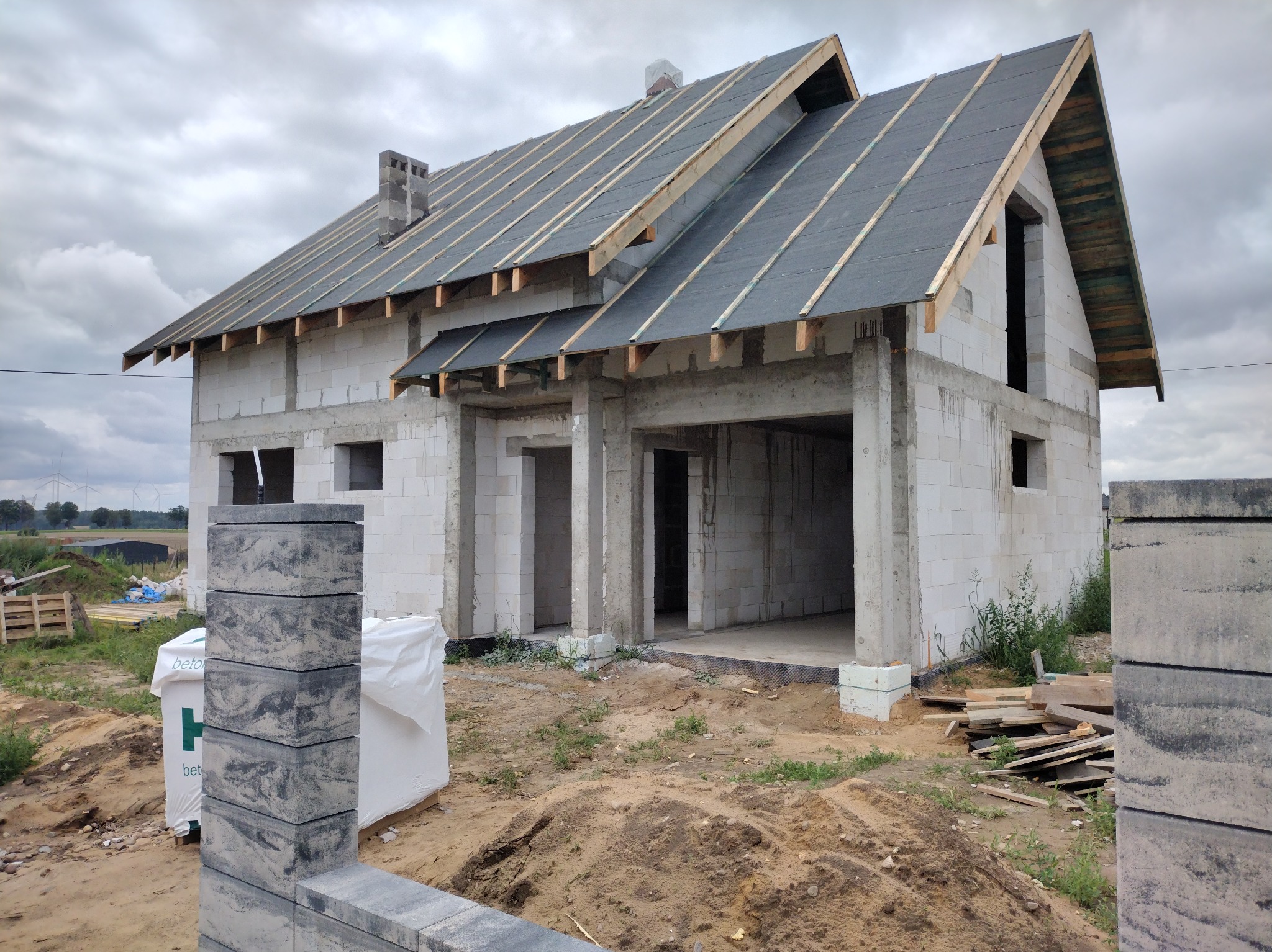 Surowy, dwukondygnacyjny dom z białego betonu komórkowego w trakcie budowy. Widoczny dach z deskowaniem i papą, komin oraz sterta bloczków betonowych na pierwszym planie.