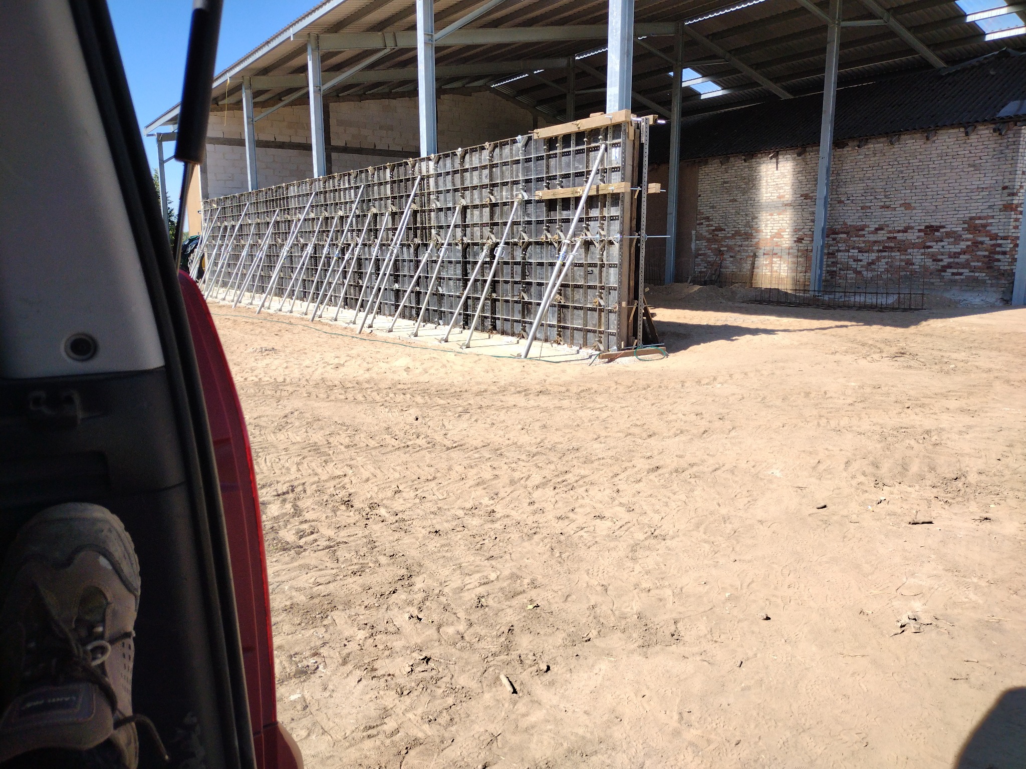 Szczegółowe ujęcie szalunku fundamentowego z metalowymi podporami na piaszczystym terenie budowy, widoczne zza otwartych drzwi samochodu.