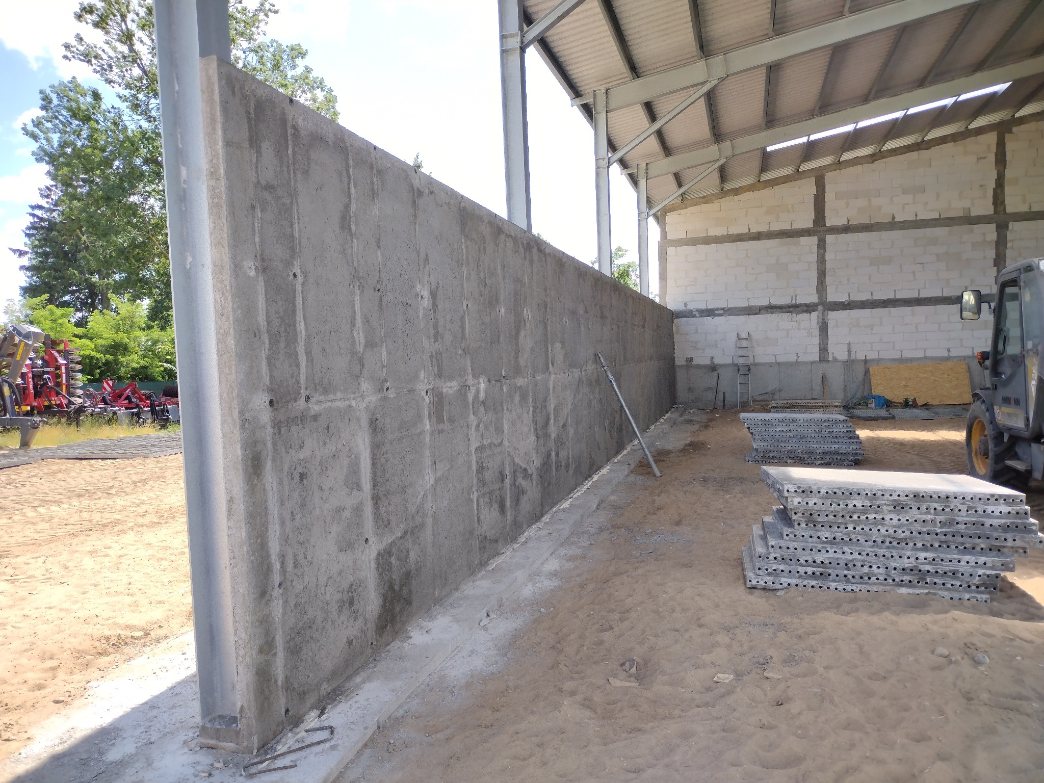 Wysoka, surowa ściana fundamentowa z betonu, z widocznymi śladami szalunku, wewnątrz hali z konstrukcją stalową i stertą płyt szalunkowych. Na zewnątrz sprzęt rolniczy.