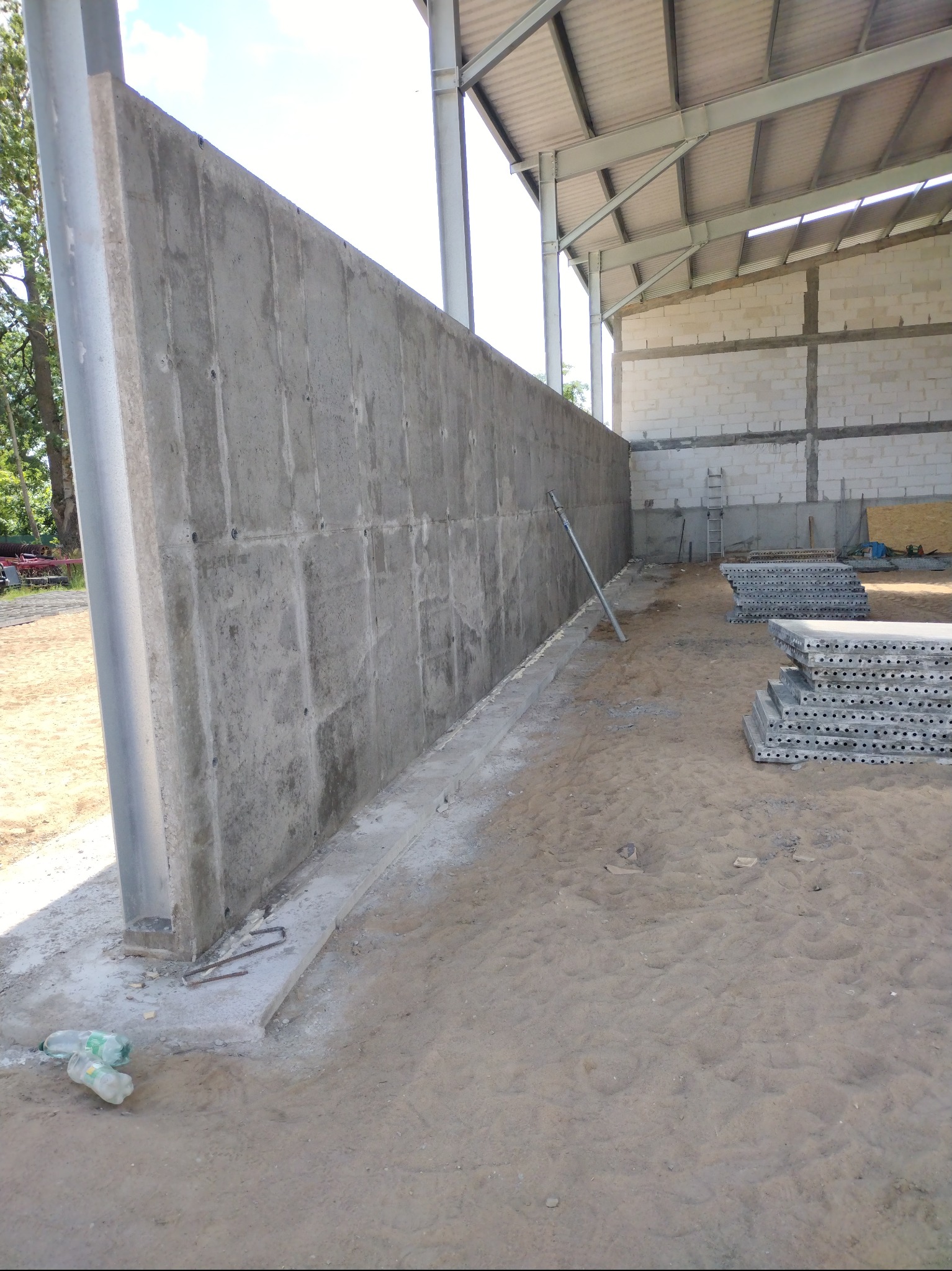 Wysoka, szara, betonowa ściana wewnątrz budynku w trakcie budowy. Widoczne metalowe elementy konstrukcyjne i stosy szalunków. Na ziemi piasek i butelki po wodzie.
