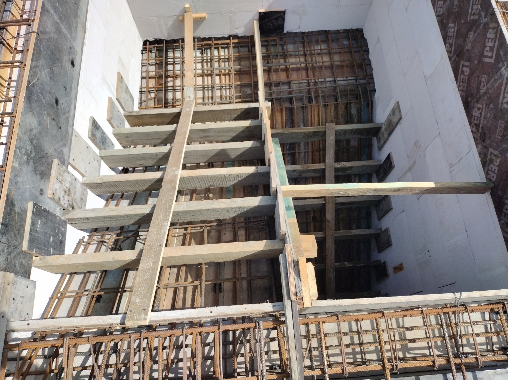 Zbrojenie schodów w trakcie budowy, widoczne szalunki i stalowe pręty. Białe ściany z bloczków betonowych w tle. Ujęcie z góry, oświetlenie naturalne.