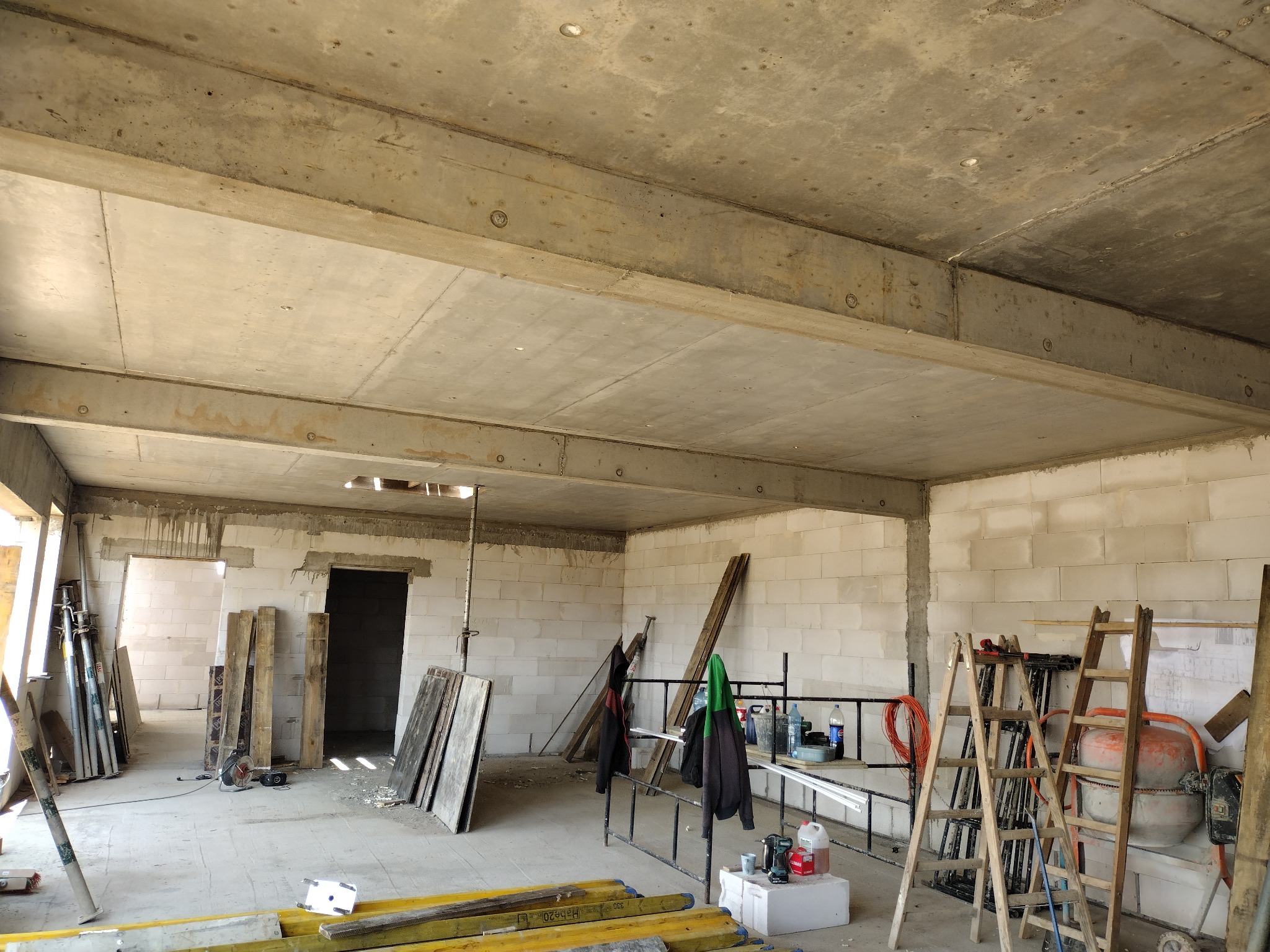 Surowy stan domu w budowie: ściany z betonu komórkowego, betonowy strop z widocznymi belkami, rusztowania i drabiny wewnątrz. Widoczne narzędzia i materiały budowlane.
