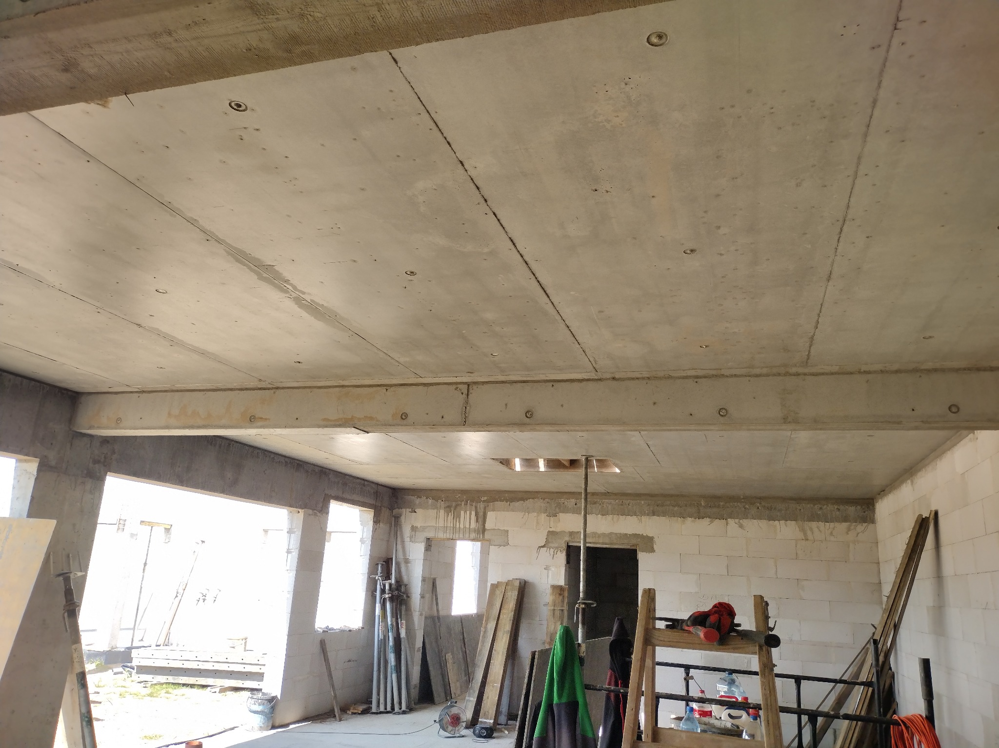 Surowy betonowy strop w nowym budynku z pustaków, widoczne otwory i belki nośne, przygotowany do dalszych prac wykończeniowych. W tle okna i materiały budowlane.