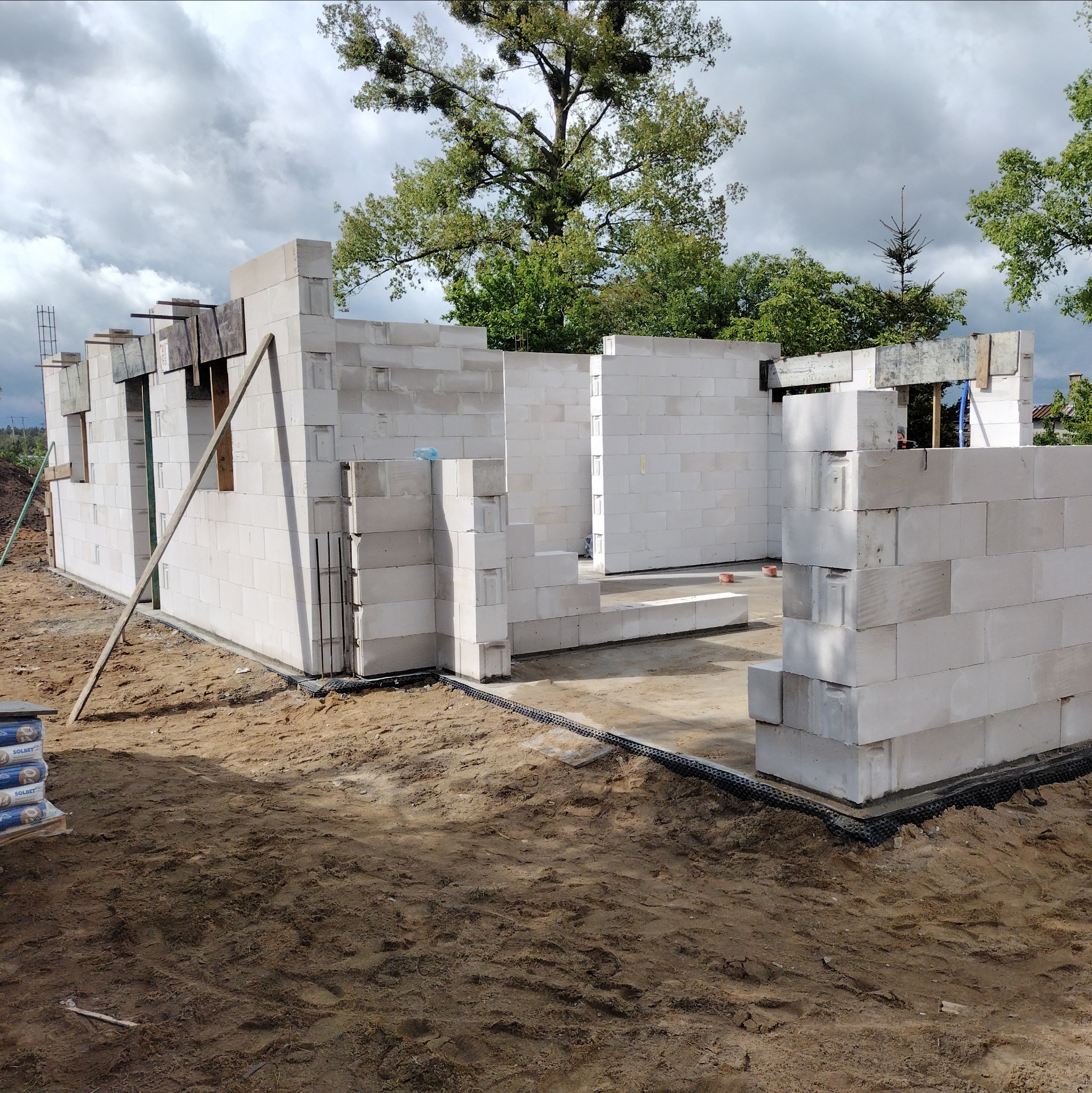 Wznoszenie ścian parteru domu z betonu komórkowego. Widoczne zbrojenia i podkłady pod okna. W tle drzewa i pochmurne niebo.