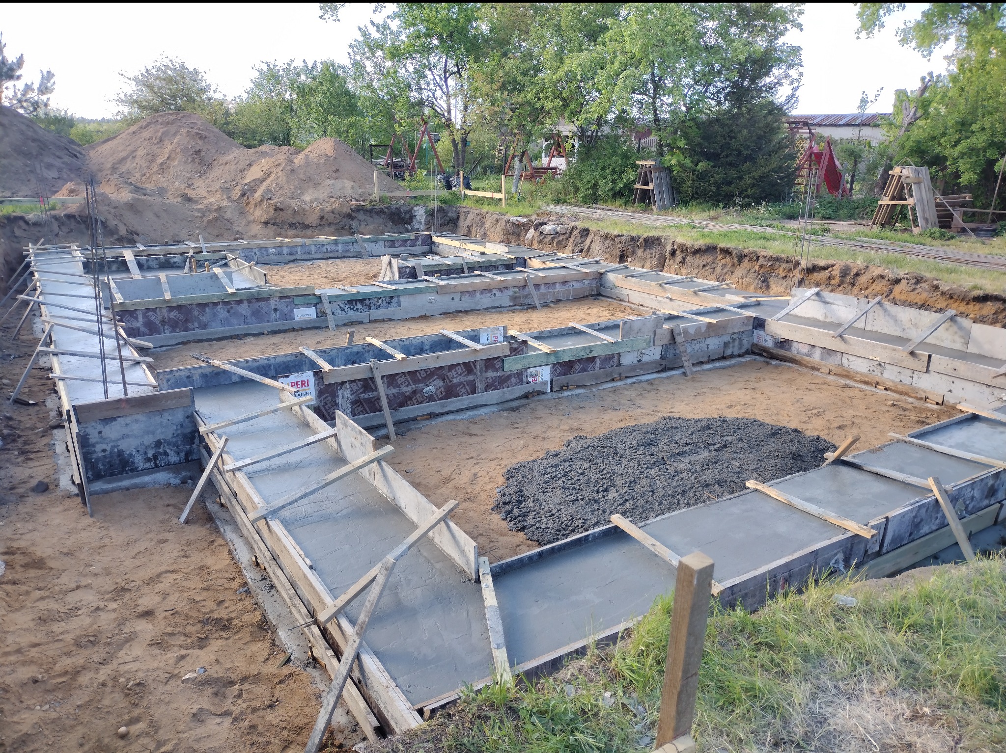 Zalane betonem szalunki fundamentowe na piaszczystym podłożu, widoczne zbrojenia i drewniane podpory. W tle hałda ziemi i zieleń drzew.