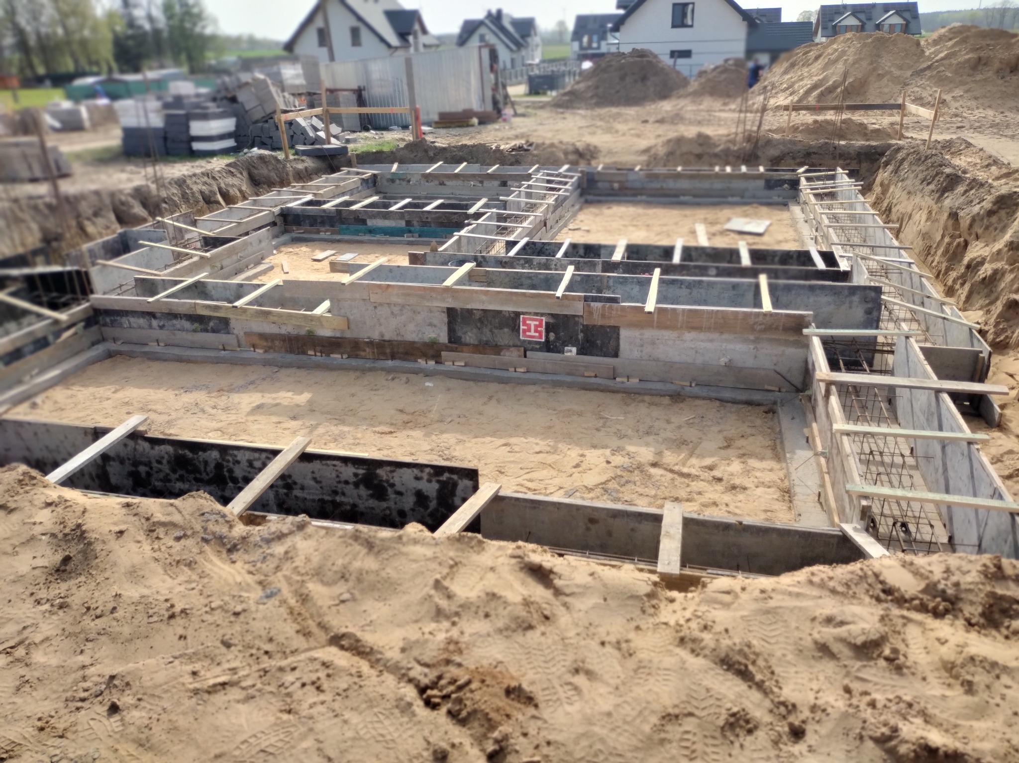Zalane fundamenty domu jednorodzinnego z widocznym zbrojeniem i szalunkiem, teren budowy z hałdami piasku w tle.