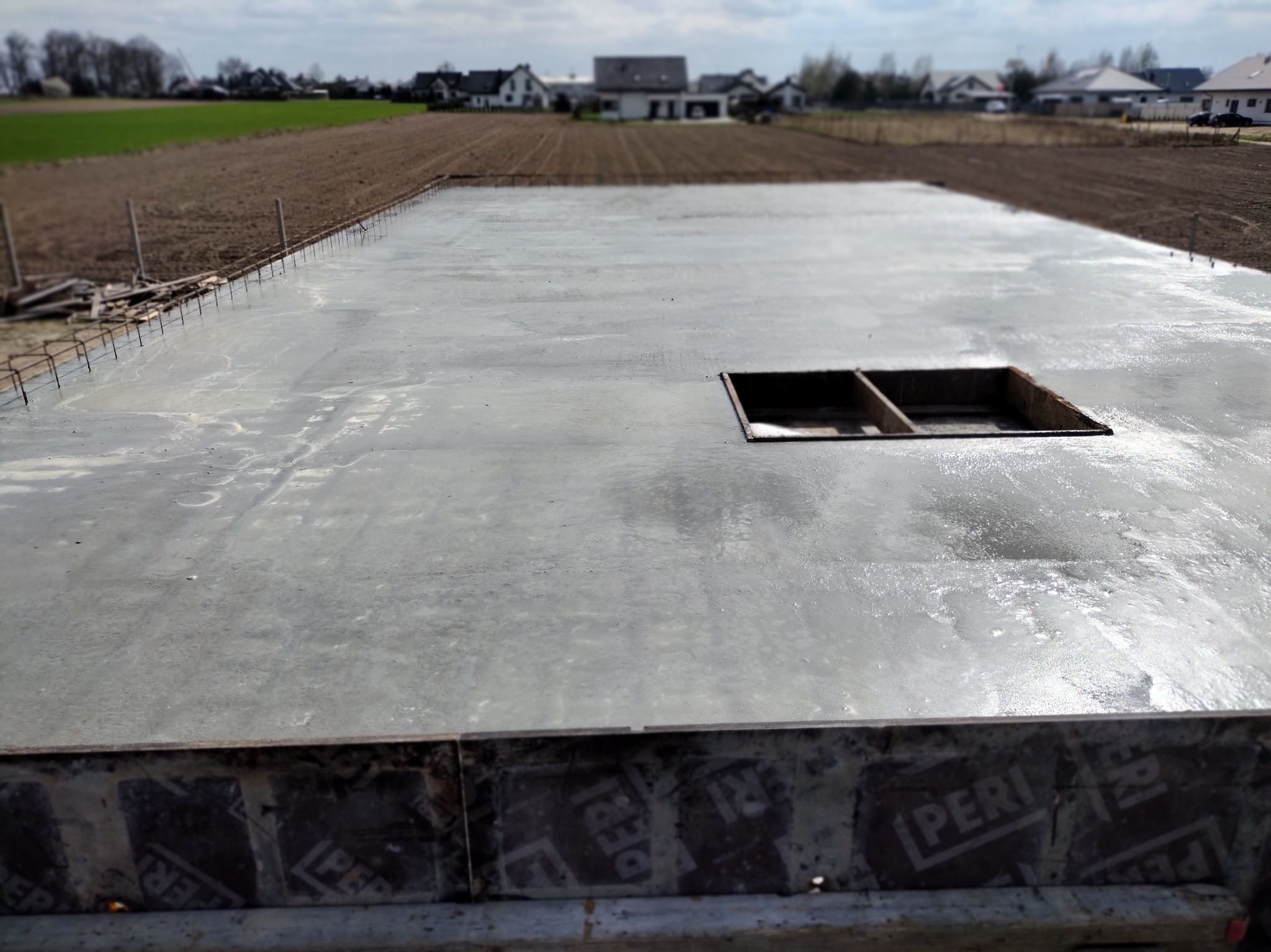 Świeżo wylana, gładka płyta fundamentowa z otworem rewizyjnym, widok z góry na tle pól i zabudowań. Widoczne zbrojenia i szalunki.