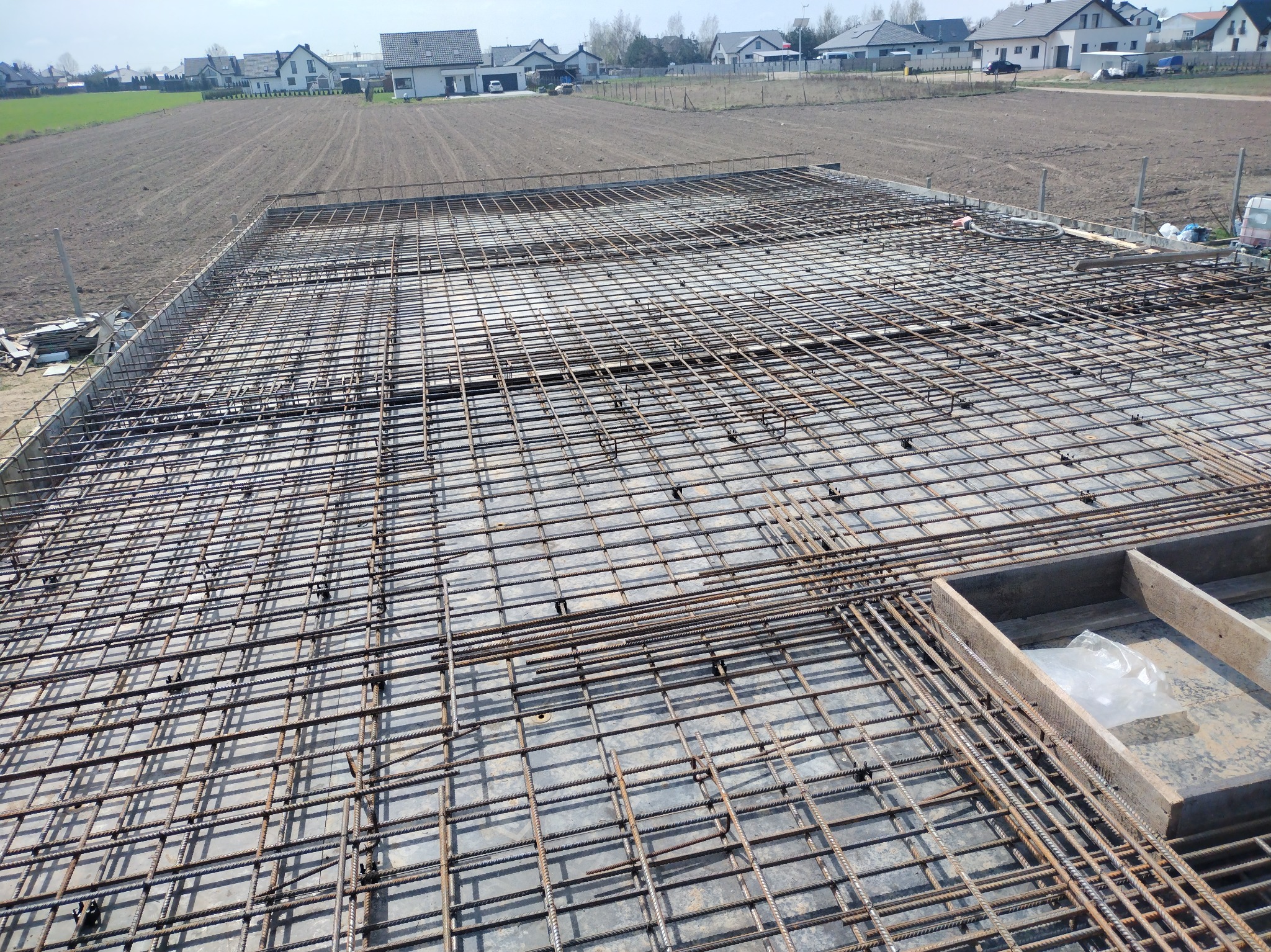 Siatka zbrojeniowa na przygotowanym fundamencie pod budowę domu jednorodzinnego. Widok z góry na konstrukcję z prętów stalowych, w tle pole i zabudowa.