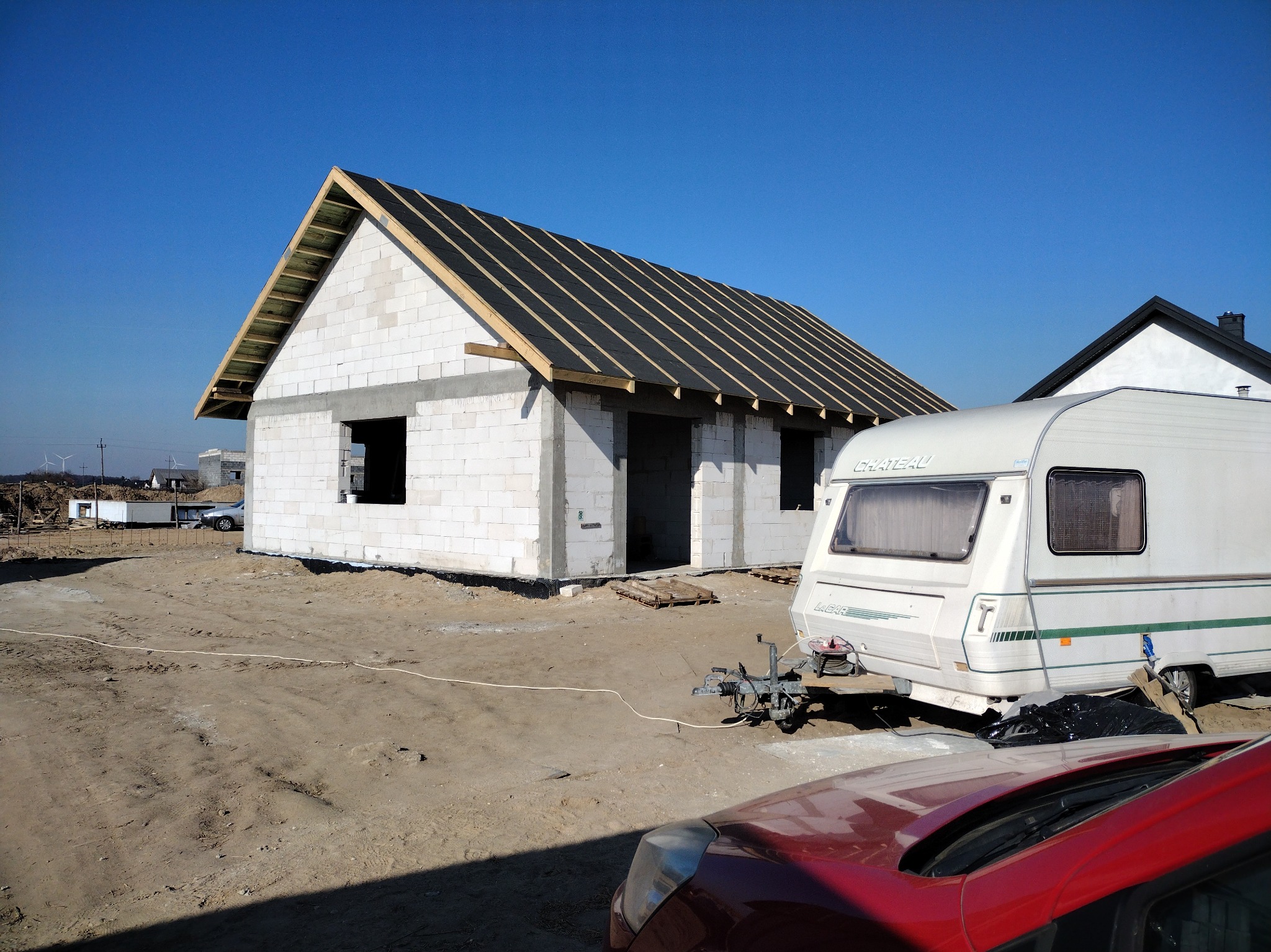 Surowy stan domu z białego bloczka, z widoczną konstrukcją dachu i czarną membraną, na tle błękitnego nieba. Przyczepa kempingowa i fragment czerwonego auta na pierwszym planie.