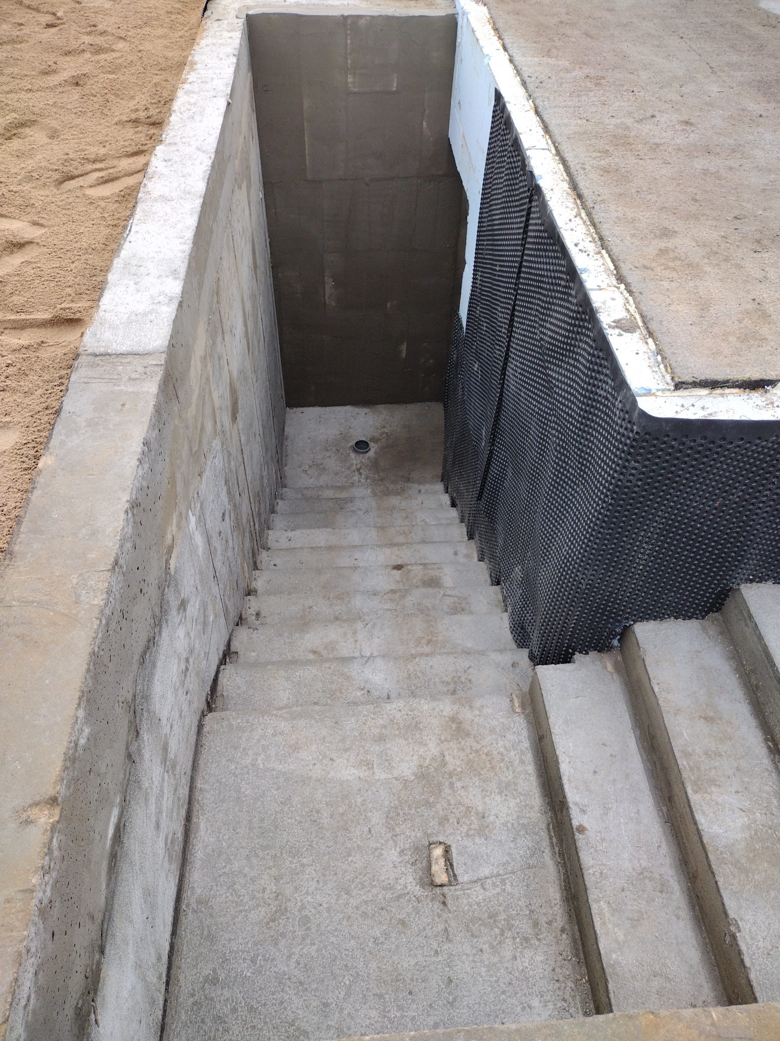 Betonowe schody do piwnicy z izolacją przeciwwilgociową z folii kubełkowej, widok z góry. Elementy konstrukcyjne w surowym stanie, piasek obok.