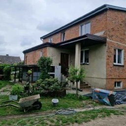 Dom Niskoemisyjny - Dwupiętrowy dom z cegły z widocznym ociepleniem części ściany, przed domem taczka, wiadro i zwinięty wąż ogrodowy, w tle widoczna skrzynka narzędziowa.