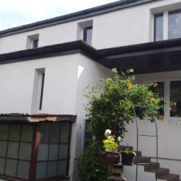 Dom Niskoemisyjny - Biały dom jednorodzinny po termomodernizacji, widoczna nowa elewacja i okna, z zielenią w ogrodzie.