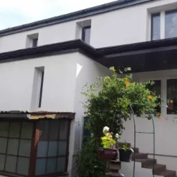 Biały dom jednorodzinny po termomodernizacji, widoczna nowa elewacja i okna, z zielenią w ogrodzie.