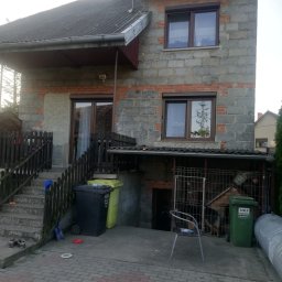 Dom Niskoemisyjny - Dwupiętrowy dom z szarego pustaka z ceglanymi akcentami, z widocznymi oknami z brązowymi ramami, schodami prowadzącymi do wejścia i różnokolorowymi koszami na śmieci przed budynkiem.