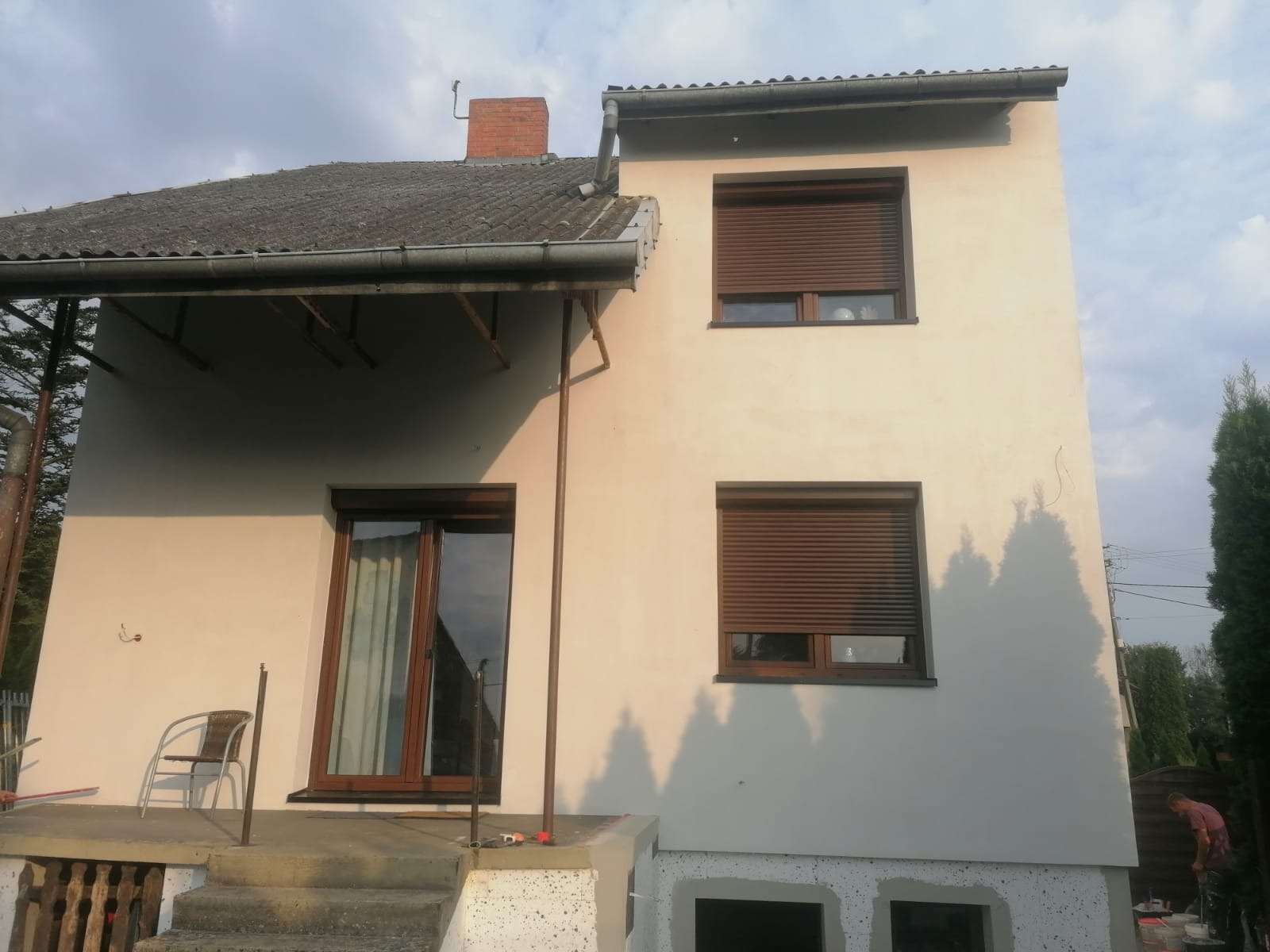 Dwupiętrowy dom z brązowymi roletami i oknami, po ociepleniu i pomalowaniu elewacji na kolor kremowy, z widocznymi rusztowaniami i pracownikiem malującym cokół budynku.