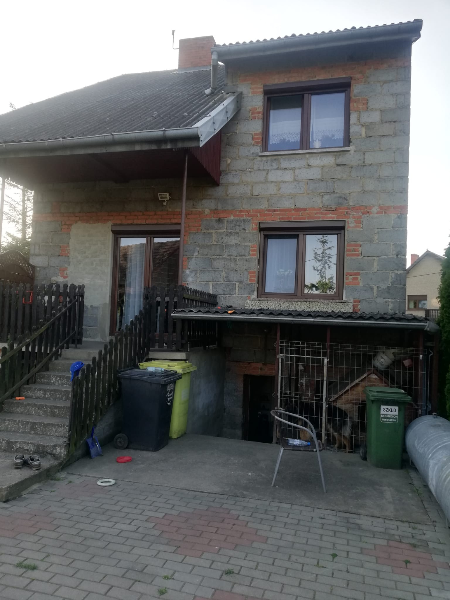 Dwupiętrowy dom z szarego pustaka z ceglanymi akcentami, z widocznymi oknami z brązowymi ramami, schodami prowadzącymi do wejścia i różnokolorowymi koszami na śmieci przed budynkiem.