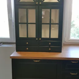 Ciemnozielona szafka kuchenna z przeszklonymi drzwiczkami i złotymi gałkami, stojąca na blacie z jasnego drewna, z widocznymi dwoma oknami w tle.
