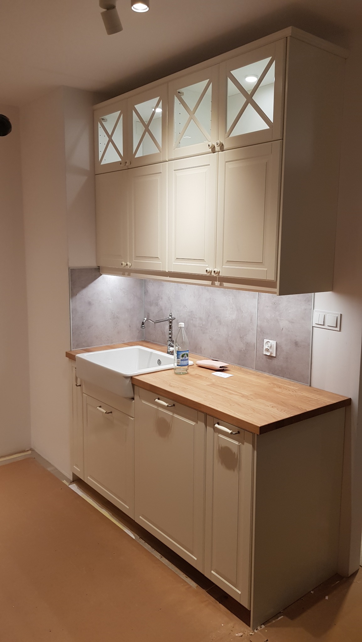 Jasna, klasyczna zabudowa kuchenna z górnymi szafkami z przeszkleniami w kształcie krzyży, drewniany blat, ceramiczny zlew typu farmhouse oraz szara ściana nad blatem.