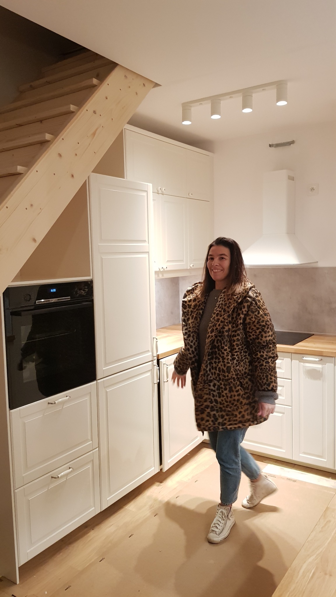 Biała zabudowa kuchenna z piekarnikiem pod schodami, kobieta w kurtce w panterkę w nowym wnętrzu, podłoga zabezpieczona kartonem.