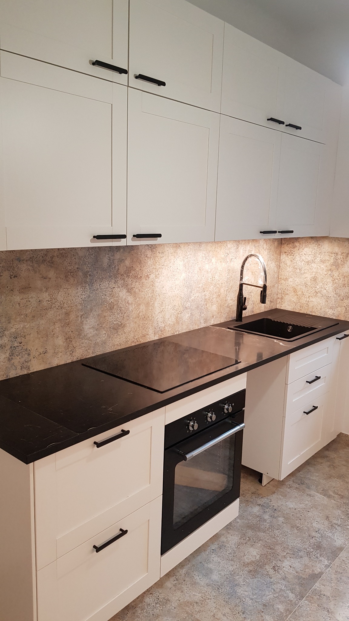 Białe szafki kuchenne z czarnymi uchwytami i blatem, czarny zlew i bateria, płyta indukcyjna, piekarnik z widocznymi pokrętłami, ściana nad blatem wykończona płytkami imitującymi kamień.