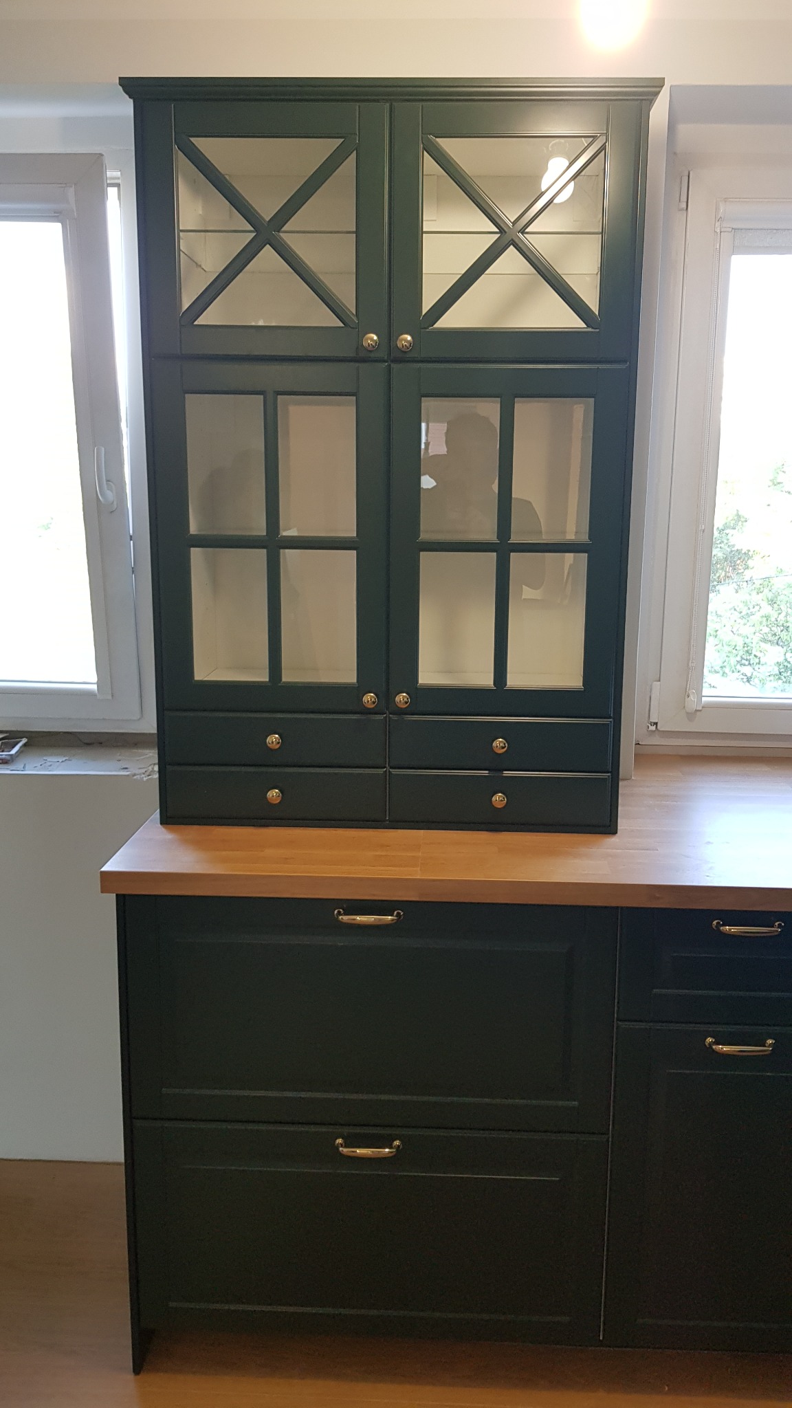 Ciemnozielona szafka kuchenna z przeszklonymi drzwiczkami i złotymi gałkami, stojąca na blacie z jasnego drewna, z widocznymi dwoma oknami w tle.