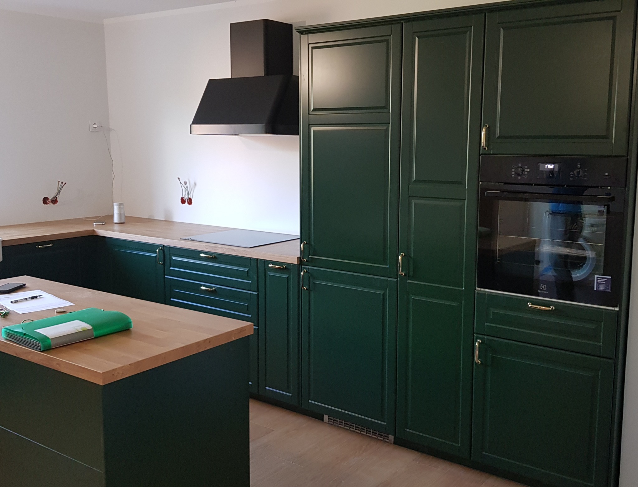 Nowoczesna kuchnia z zielonymi meblami na wymiar, drewnianym blatem i czarnym okapem, widoczne niedokończone instalacje elektryczne na ścianie.