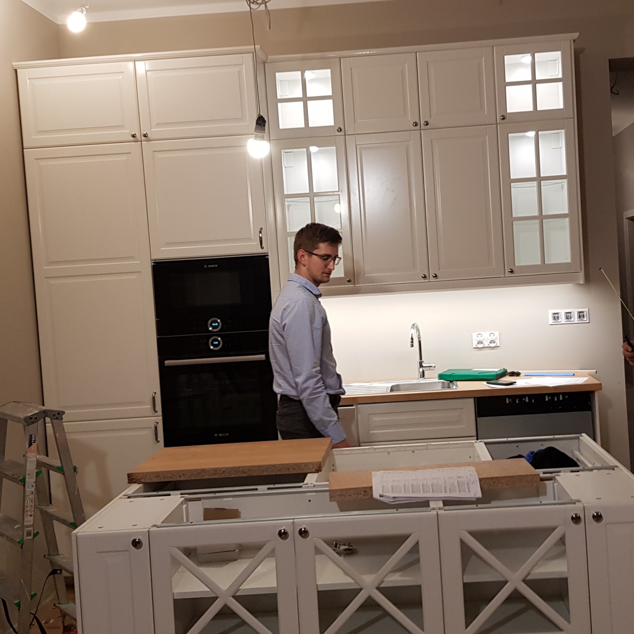 Montaż białej zabudowy kuchennej z oświetleniem podszafkowym; widoczny mężczyzna w koszuli, drabina i częściowo zmontowana wyspa kuchenna z dekoracyjnymi krzyżami na frontach.