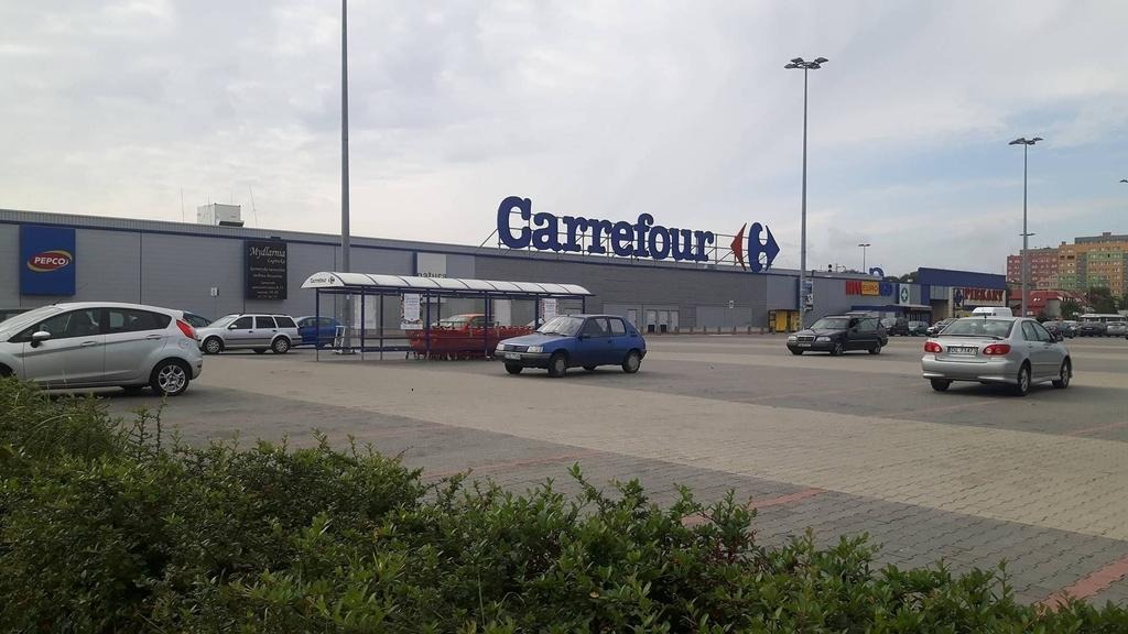 Parking przed dużym budynkiem handlowym Carrefour z widocznymi samochodami osobowymi, wózki sklepowe pod zadaszeniem i budynki w tle.
