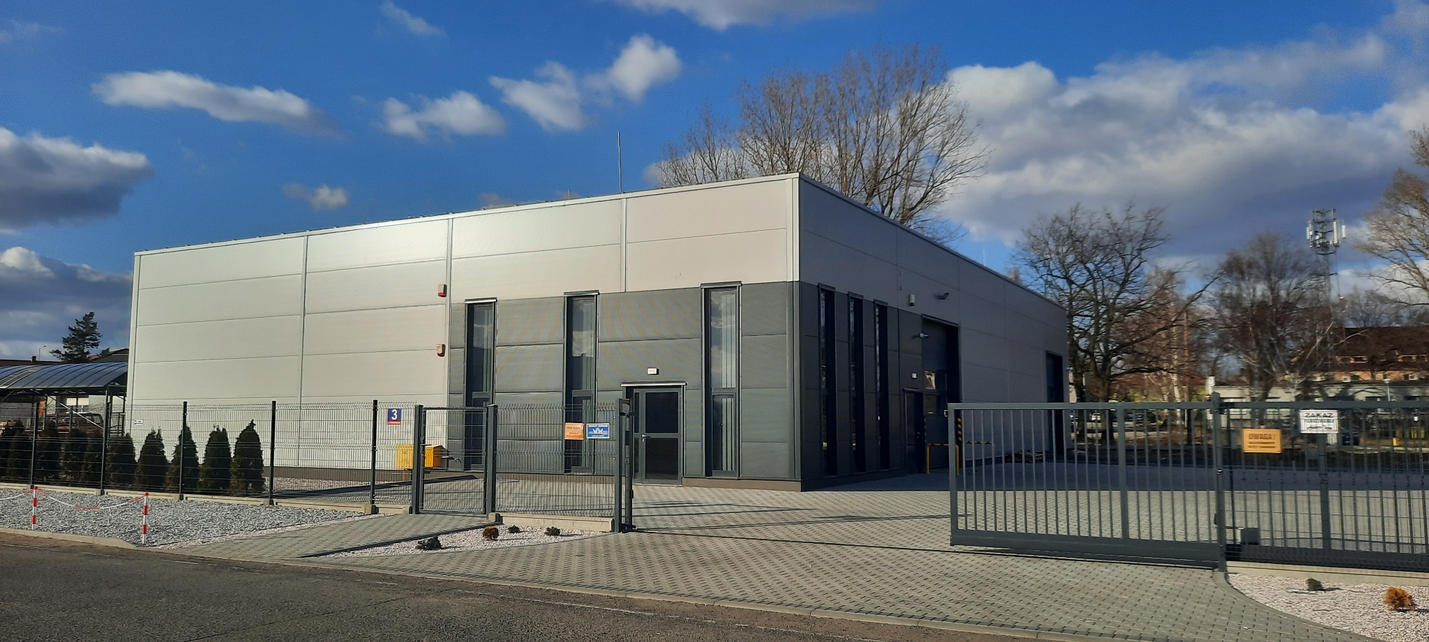 Nowoczesny budynek przemysłowy z szarego i białego panelu, ogrodzony metalowym płotem z bramą wjazdową, widoczne drzewa i błękitne niebo z chmurami.