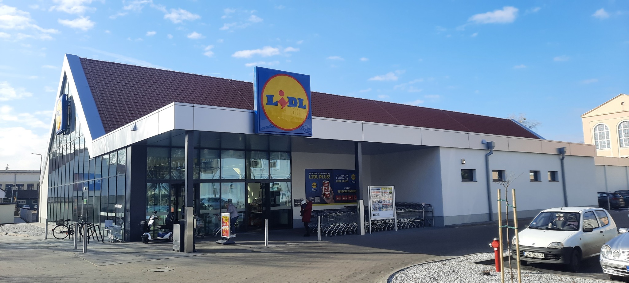Nowoczesny budynek sklepu Lidl z charakterystycznym żółto-niebieskim logo, widoczny w słoneczny dzień z parkingu z wózkami i zaparkowanymi samochodami.