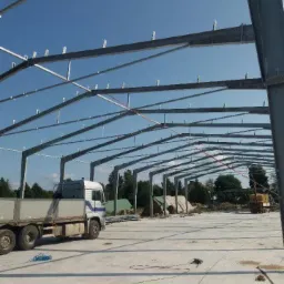 Szkielet konstrukcji stalowej hali w trakcie budowy, widoczny niebieski niebo, ciężarówka i materiały budowlane na placu budowy.