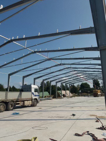 Szkielet konstrukcji stalowej hali w trakcie budowy, widoczny niebieski niebo, ciężarówka i materiały budowlane na placu budowy.