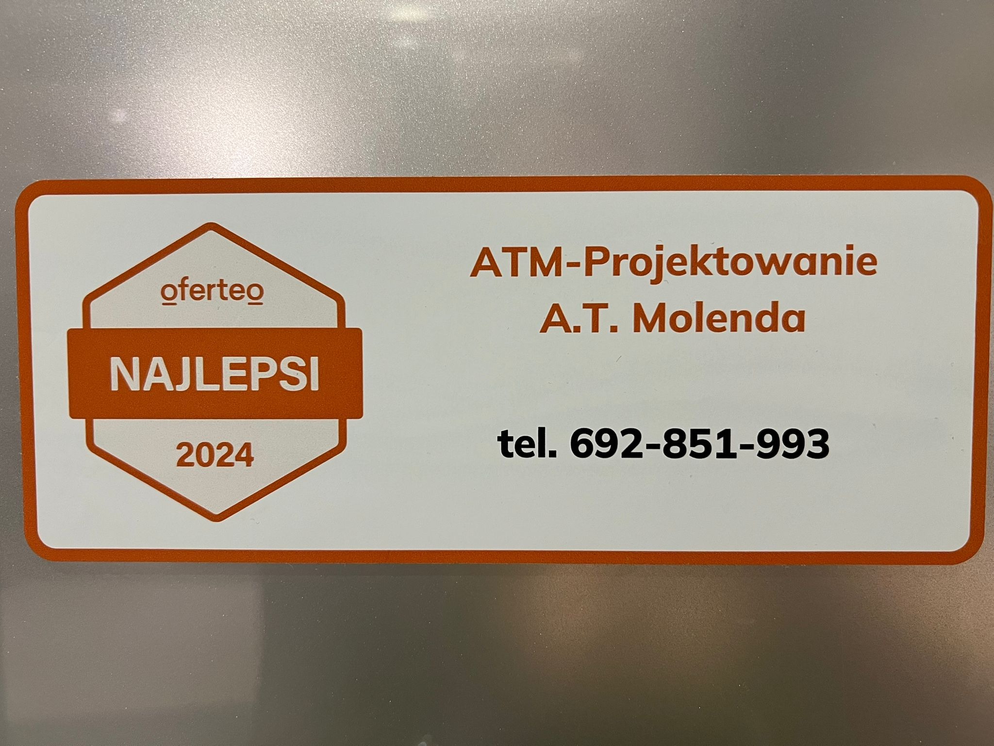 Naklejka z logo Oferteo 'Najlepsi 2024' oraz danymi kontaktowymi firmy ATM-Projektowanie A.T. Molenda i numerem telefonu na srebrnym tle.