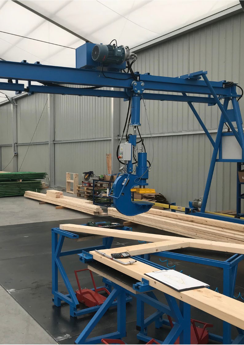 Niebieska suwnica z chwytakiem hydraulicznym podnosi drewniane elementy w hali produkcyjnej. Na pierwszym planie stół roboczy z dokumentacją techniczną i kalkulatorem.