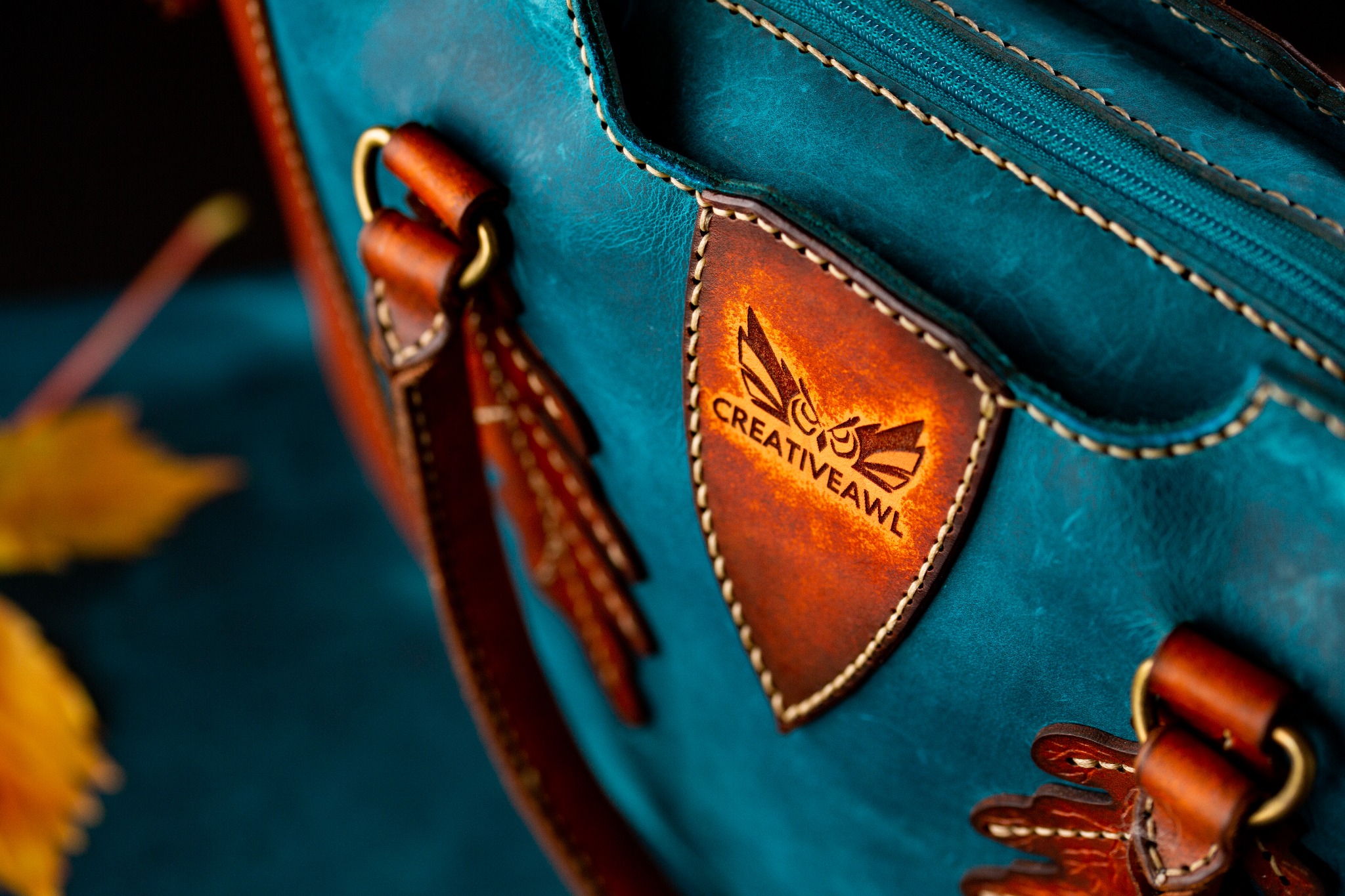 Turkusowa skórzana torba z brązowymi detalami, w tym logo 'Creativeawl' na naszywce i skórzane uchwyty, widoczne szwy i zamek błyskawiczny, liść w tle.