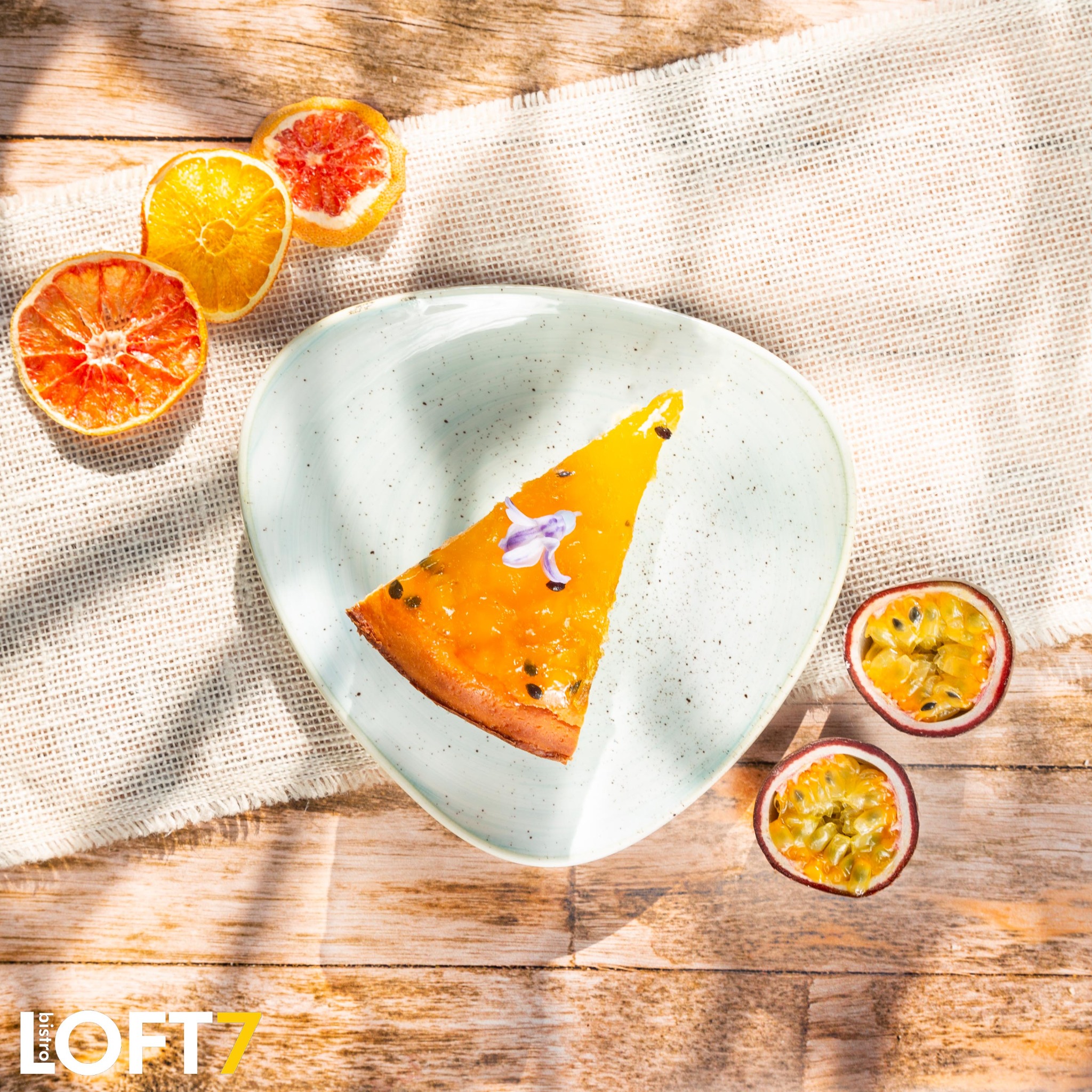 Kawałek ciasta z marakują udekorowany kwiatem na trójkątnym talerzu, otoczony suszonymi i świeżymi cytrusami na lnianej ściereczce i drewnianym stole w jasnym świetle.