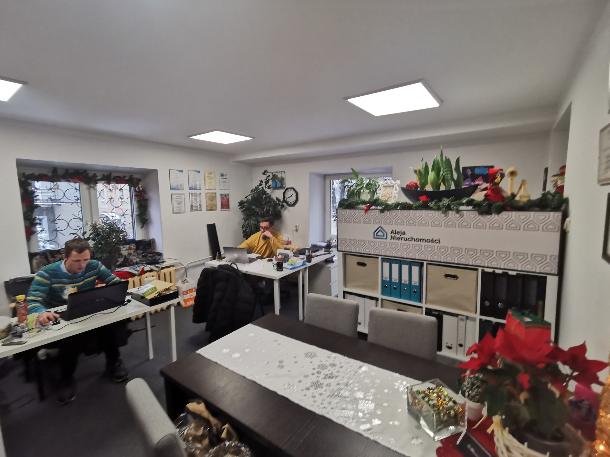 Wnętrze biura nieruchomości z dwoma pracownikami przy biurkach, jeden w niebiesko-zielonym swetrze korzysta z laptopa, drugi w żółtej koszuli rozmawia przez telefon, w tle regał z logo firmy...