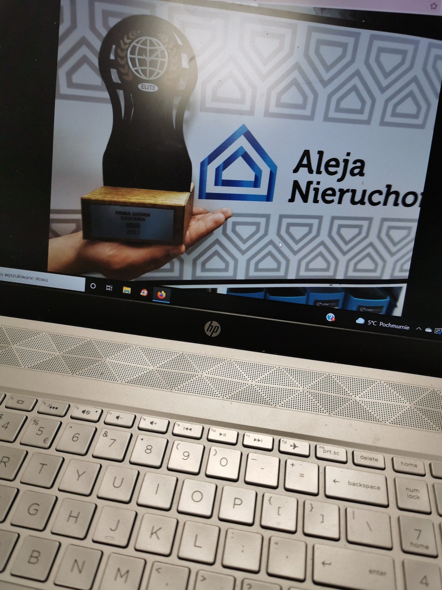 Zdjęcie ekranu laptopa z otwartą stroną firmy Aleja Nieruchomości prezentującą nagrodę 'Firma Godna Zaufania 2021' oraz statuetkę Elite, widoczna klawiatura laptopa.