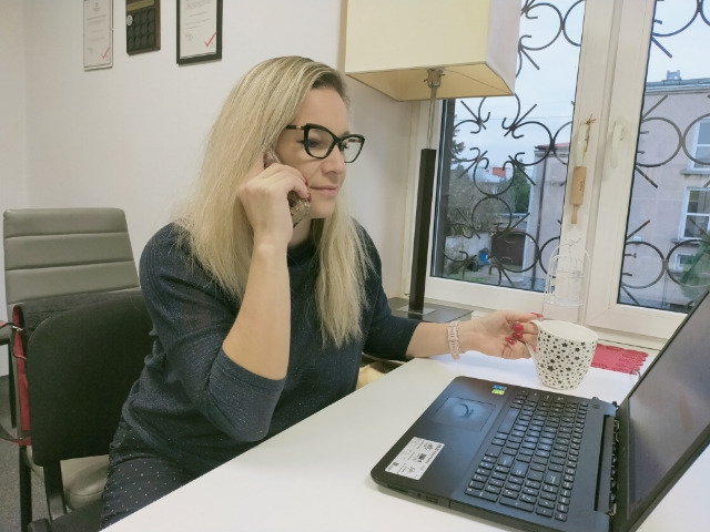 Blondynka w okularach rozmawia przez telefon, trzymając filiżankę w dłoni, obok laptopa przy biurku w biurze z oknem i ozdobną lampą.