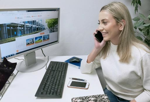 Kobieta z blond włosami rozmawia przez telefon przy biurku, na którym stoi monitor z otwartą stroną internetową agencji nieruchomości, klawiatura, myszka, telefon i długopis. W tle widać czarnego...