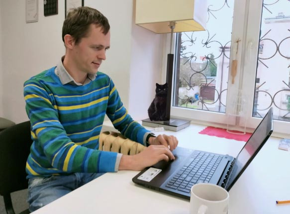 Mężczyzna w pasiastym swetrze pracuje na laptopie przy biurku z widokiem na okno, gdzie stoi lampka z czarną figurką kota.