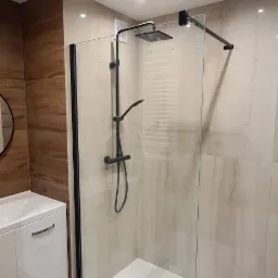 Nowoczesna kabina prysznicowa walk-in z czarnymi detalami, zamontowana na białym brodziku, z odpływem liniowym, na tle płytek imitujących drewno i beżowego marmuru, oświetlona punktowymi lampami...
