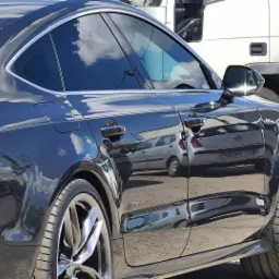 Błyszczący, czarny samochód Audi A7 z przyciemnionymi szybami bocznymi, odbijający chmury i otoczenie w lakierze. Widoczne chromowane felgi i klamki. W tle zaparkowany biały samochód ciężarowy.