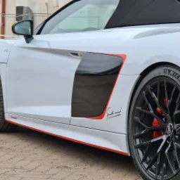 Białe Audi R8 Spyder z czarnymi felgami i czerwonymi akcentami na karoserii, widoczne z boku na tle kostki brukowej.