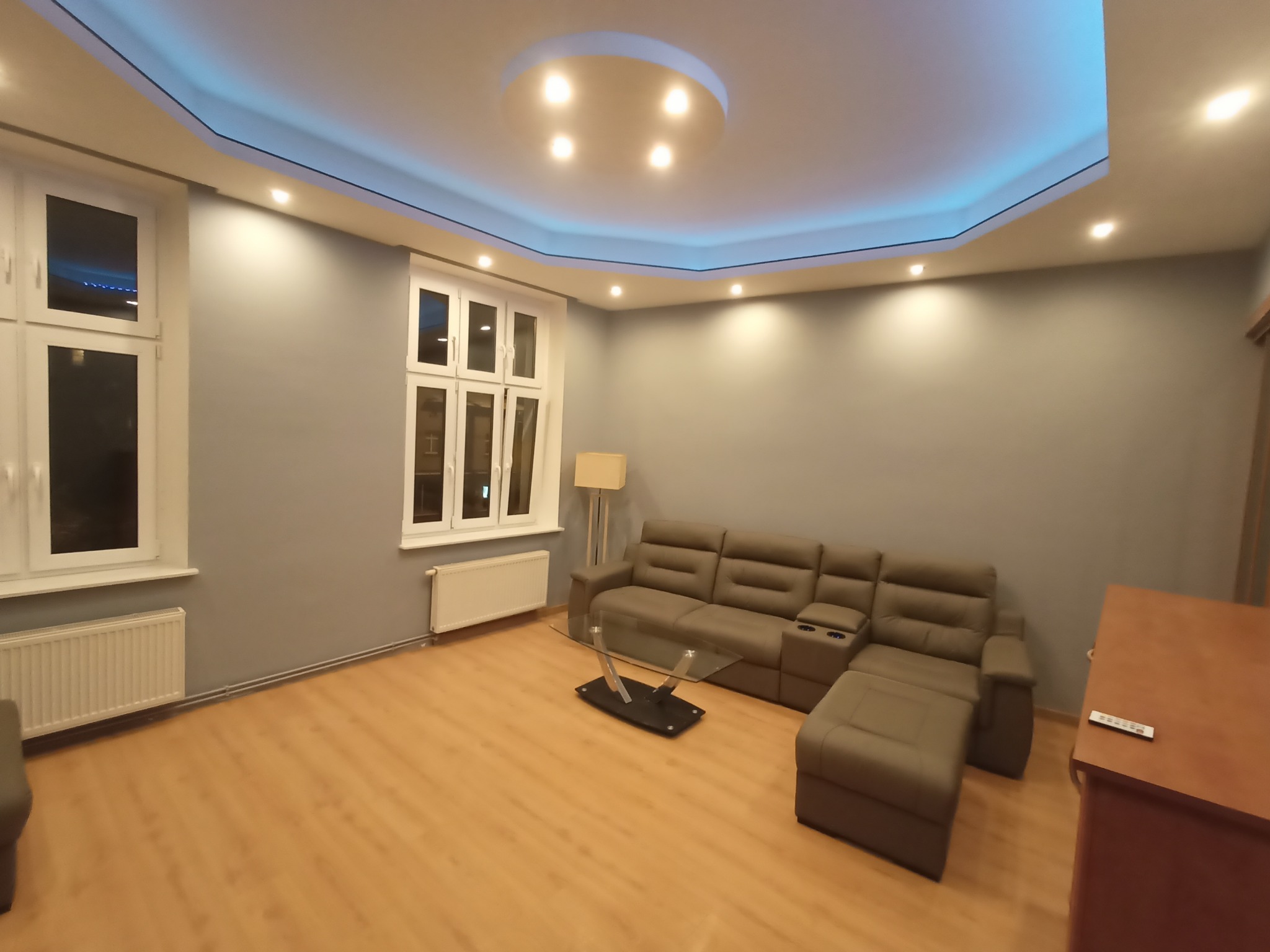 Salon po remoncie: szare ściany, drewniana podłoga, narożnik z funkcją relaks, szklany stolik. Sufit z podświetleniem LED. Dwa okna z białymi ramami.