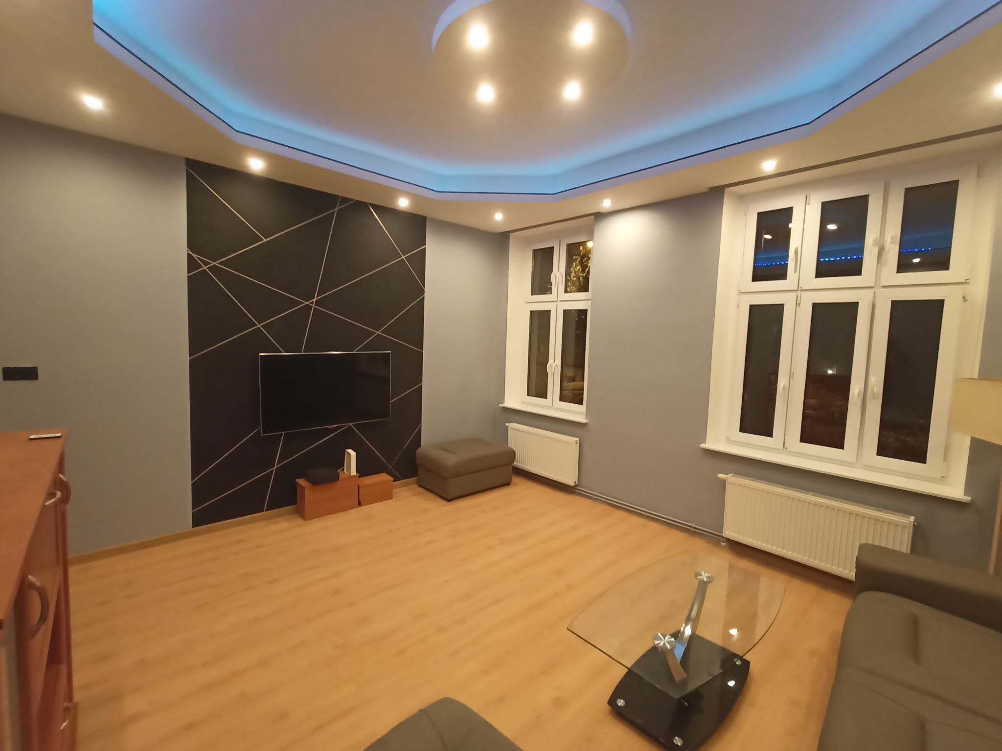 Nowoczesny salon z szarą ścianą, geometrycznym wzorem za telewizorem i podświetlanym sufitem. Parkiet na podłodze, okna z białymi ramami, grzejniki.