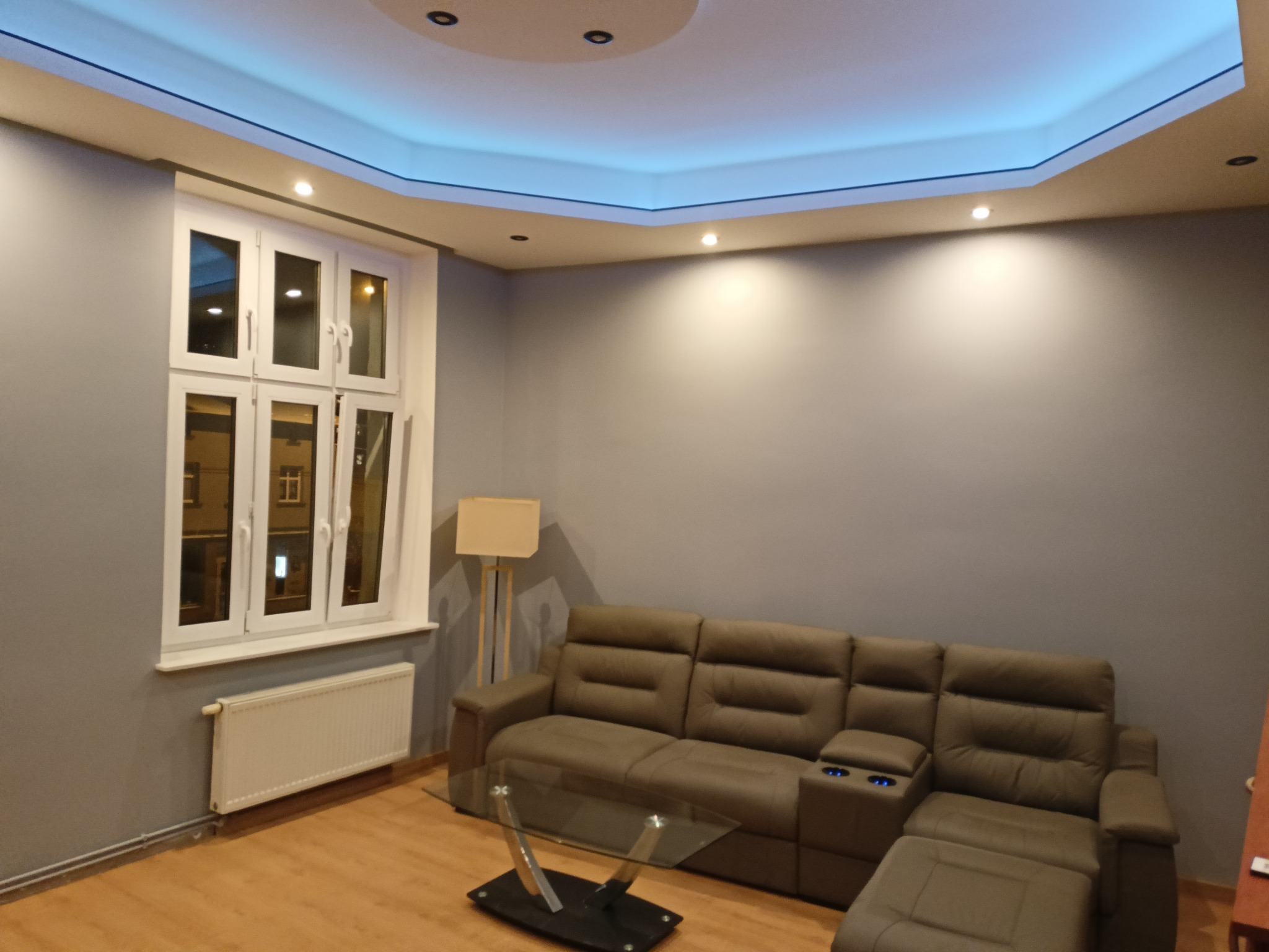 Stylowy salon z szarą ścianą, brązową sofą i szklanym stolikiem. Białe okno z widokiem na zewnątrz. Sufit z podświetleniem LED. Nowoczesne wykończenie wnętrza.