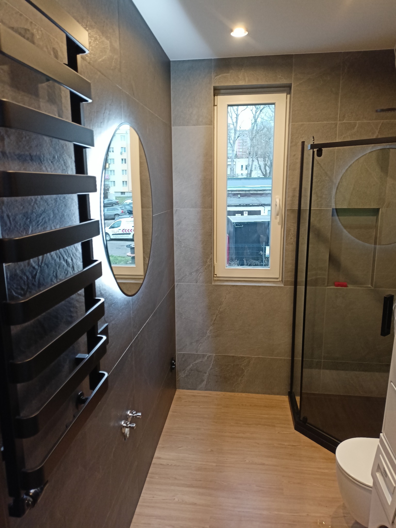 Nowoczesna łazienka z szarymi płytkami, owalnym lustrem z podświetleniem, czarnym grzejnikiem drabinkowym i kabiną prysznicową z czarnymi profilami.